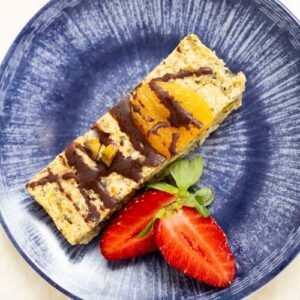 A rectangular granola bar with chocolate drizzle and fruit pieces is served on a blue plate with two strawberry halves.