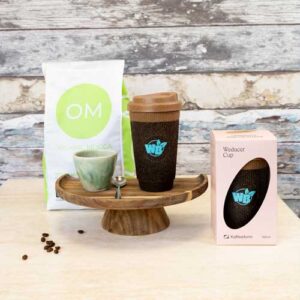 Una taza de café reutilizable, una taza de cerámica con cuchara, una bolsa de café ecológico y una taza reutilizable en caja se exponen en un soporte y una mesa de madera.