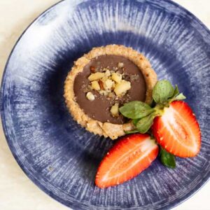 Una pequeña tarta de chocolate cubierta de nueces picadas se sirve en un plato azul con dos mitades de fresa fresca.