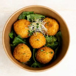 Cinco bolas empanadas y fritas se sirven en un cuenco marrón sobre un lecho de hojas verdes, coronadas con una pequeña guarnición de brotes.