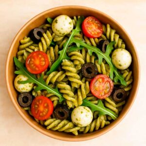 A bowl of fusilli pasta with pesto, cherry tomatoes, black olives, mozzarella balls, and arugula leaves.