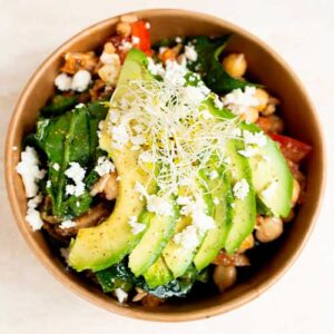A bowl filled with chickpeas, spinach, sliced avocado, crumbled cheese, sprouts, and diced vegetables.