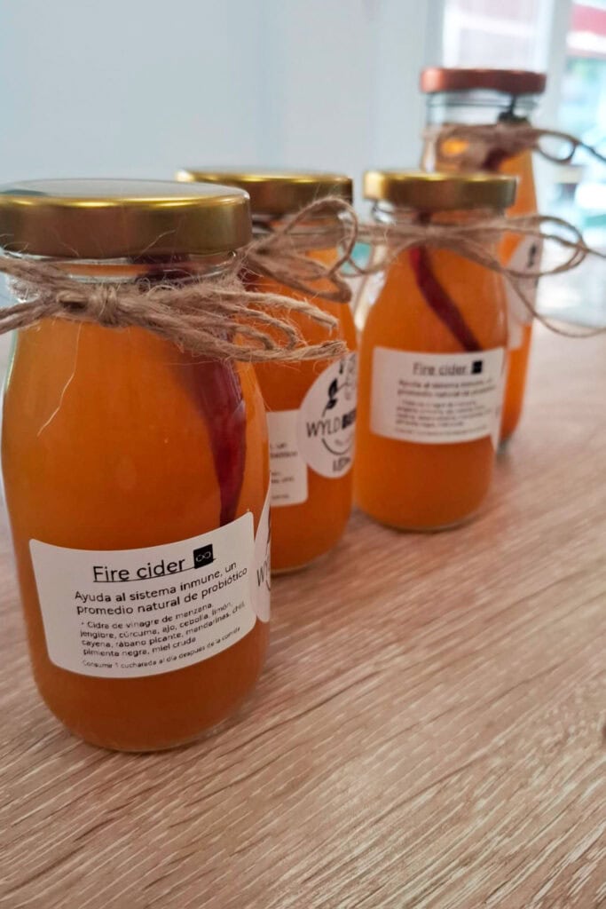 Four glass bottles filled with orange liquid labeled “Fire cider,” each sealed with a gold lid and twine, are arranged on a wooden surface.