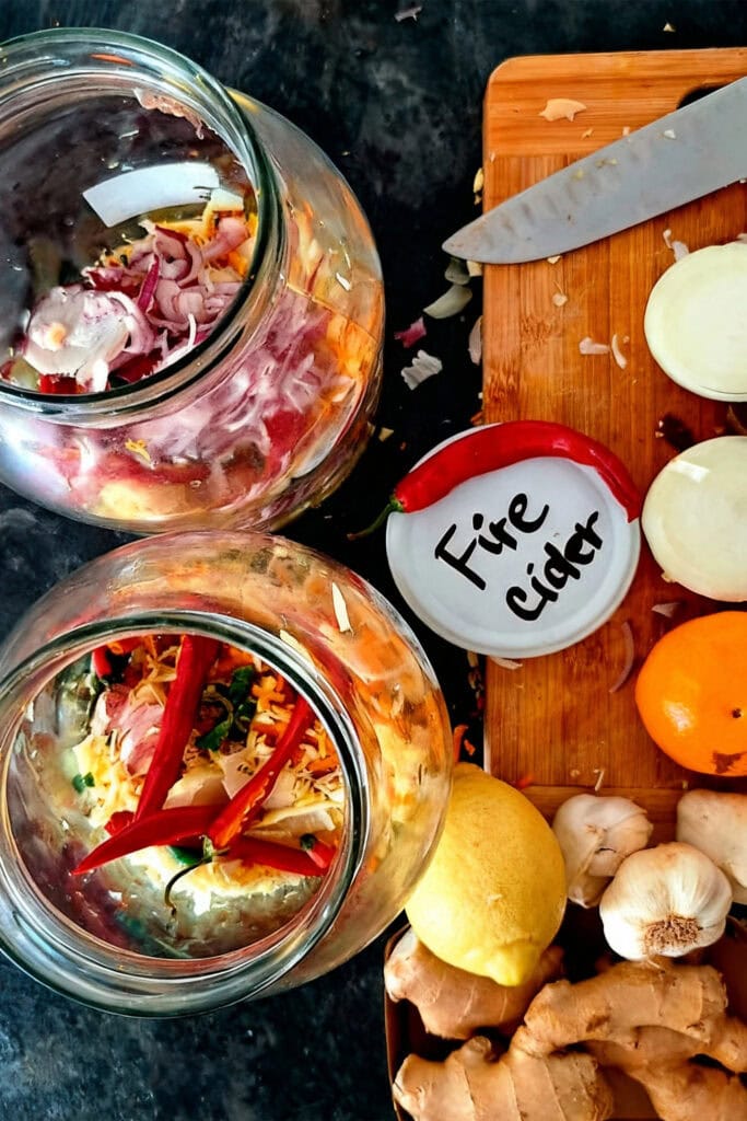 Two glass jars with sliced vegetables and spices labeled "Fire Cider" next to a cutting board with onions, citrus fruits, garlic, ginger, and a knife on a dark countertop.