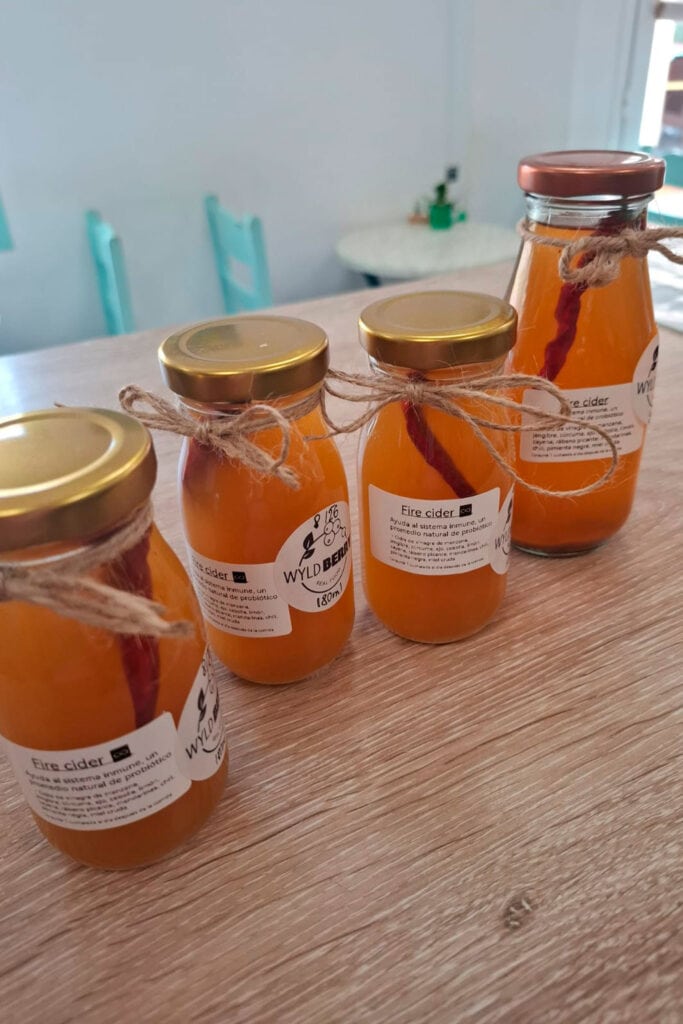 Four glass jars filled with orange liquid, labeled "Fire cider," are arranged in a row on a wooden surface. Two jars have twine tied around their necks.