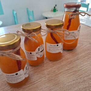 On a wooden table are displayed four glass bottles filled with orange Fire Cidre - a shot that strengthens immunity - each of them sealed with a golden cap and tied with string.