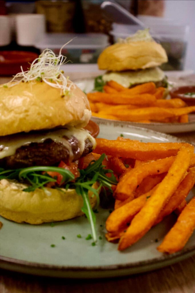 A cheeseburger with fresh greens and fries is served on a plate, with another similar meal in the background.