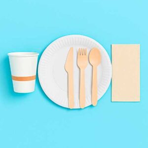 Un vaso de papel, un plato de papel blanco con un cuchillo de madera, un tenedor, una cuchara y una servilleta beige están dispuestos sobre un fondo azul.