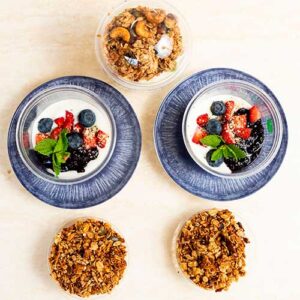 Two bowls of yogurt topped with berries, mint, and fruit preserves on blue plates, with three small bowls of granola nearby.