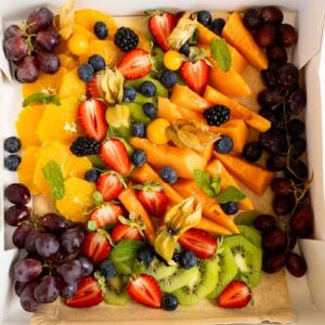 A box filled with sliced oranges, kiwi, cantaloupe, strawberries, grapes, blueberries, blackberries, physalis, and mint leaves arranged neatly on parchment paper.
