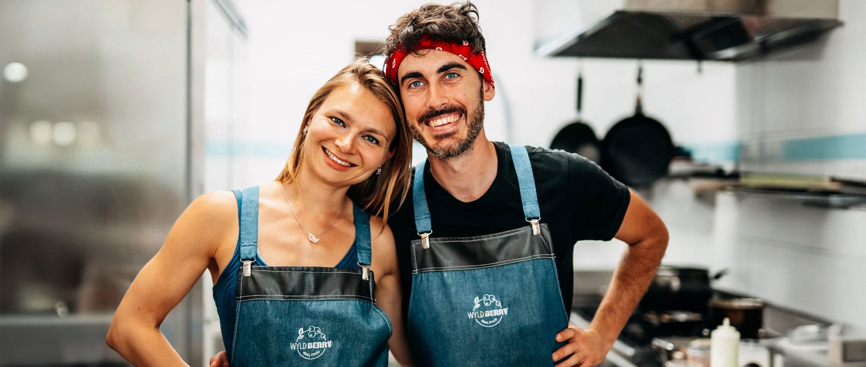 Two people in aprons stand close together in the Calvia Restaurant’s commercial kitchen, smiling at the camera.