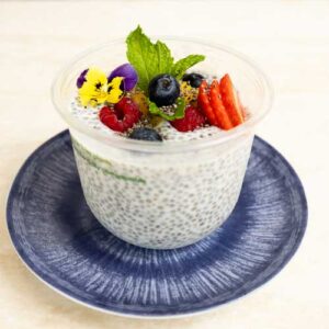 Eine Portion Veganer Chia Pudding mit Mango und roten Früchten 280g, garniert mit Erdbeeren, Heidelbeeren, Himbeeren (rote Früchte), essbaren Blüten und einem Minzblatt auf einem blauen Teller.