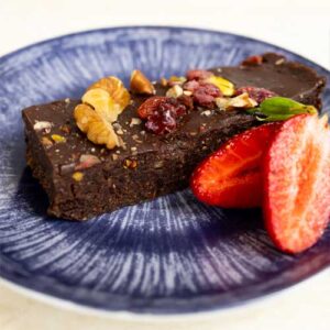 Eine Superfood-Brownie-Schnitte mit Nüssen und Trockenfrüchten, serviert mit frischen Erdbeerhälften auf einem blauen Teller.