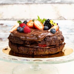Eine Rote-Bete-Schokoladen-Tarte (ganz) mit Schokoladenglasur, Erdbeeren, Himbeeren, Heidelbeeren, Brombeeren und Minzblättern auf einem gläsernen Tortenständer.