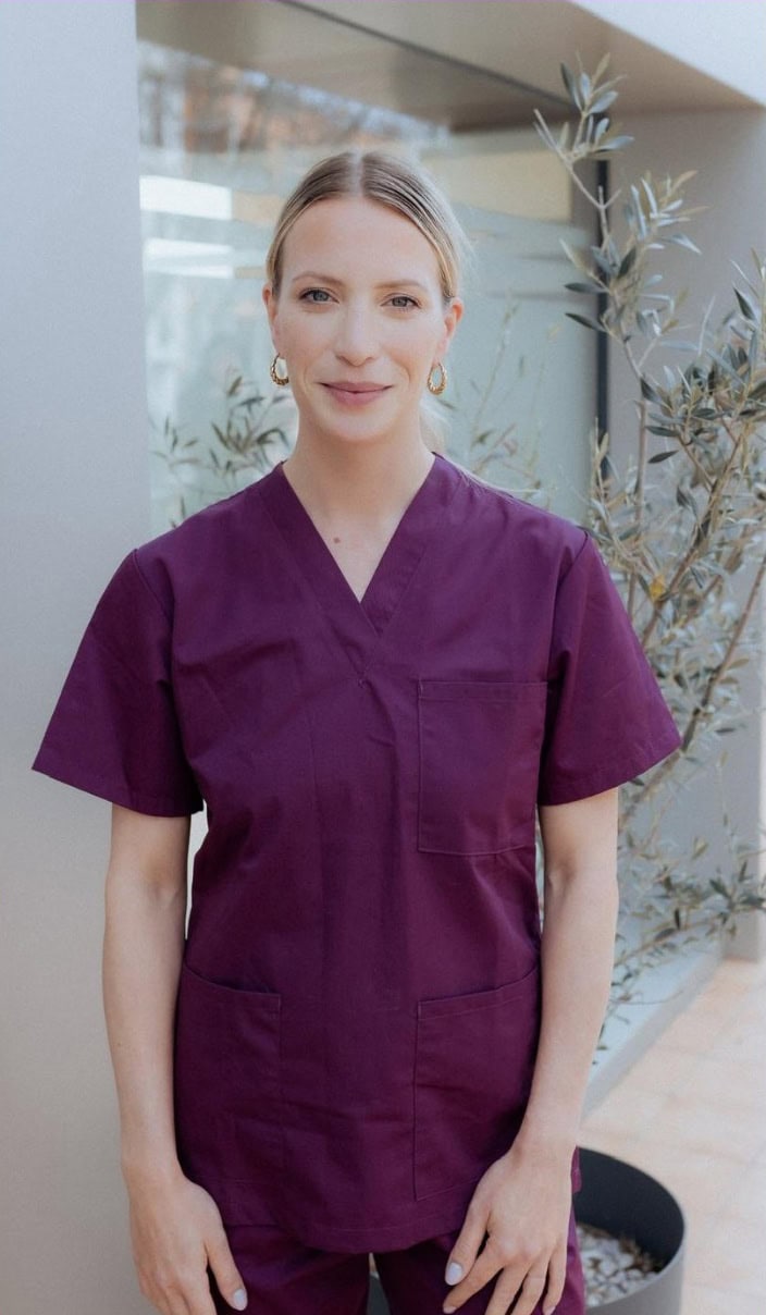 A woman wearing purple medical scrubs stands outdoors in front of a modern building and a potted plant, looking at the camera and smiling slightly—embracing better living as part of her daily routine.