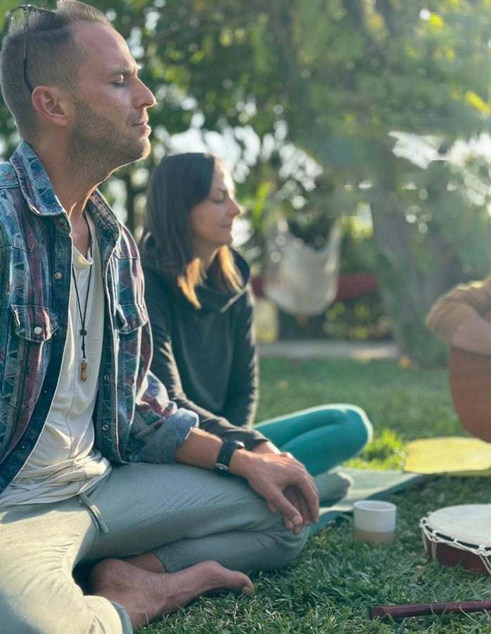 Dos personas sentadas con las piernas cruzadas en la hierba, al aire libre, con los ojos cerrados, parecen meditar. Un tambor y una copa están en el suelo. La luz del sol se filtra entre los árboles del fondo, evocando serenidad y hábitos que ayudan a vivir mejor.