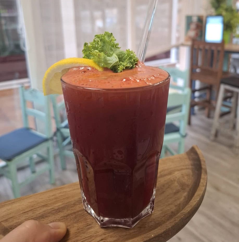 A glass of dark red smoothie garnished with a lemon slice and kale, served with a straw on a wooden tray in a café setting—perfect for those who value healthy living.
