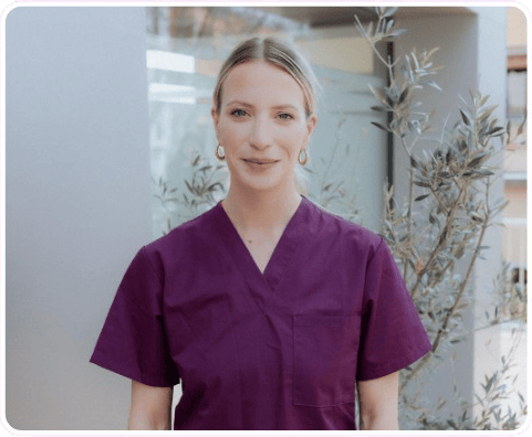A woman wearing purple medical scrubs stands outside in front of a building and some plants, embodying the spirit of longevity as she looks confidently at the camera.
