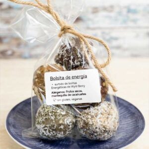 A clear plastic bag tied with twine contains assorted energy balls on a blue plate. The label indicates ingredients and allergen information in Spanish.